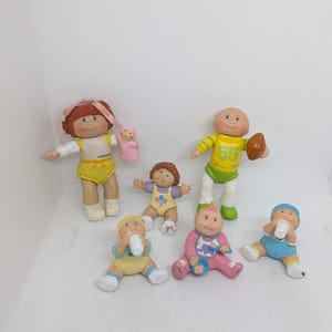 May include: A collection of vintage Cabbage Patch Kids figurines. The dolls are in various poses and outfits, including a football player, and babies with bottles. The color palette includes yellow, pink, blue, and brown.