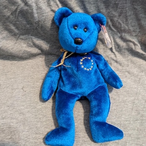 May include: A plush blue teddy bear with a black nose and small black eyes. The bear has a gold ribbon around its neck and a gold starburst design on its chest. A clear tag is attached to its ear.