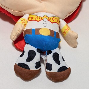May include: A soft toy doll of a character from the Toy Story franchise. The doll has a white shirt with a yellow yoke, blue trousers, and cow-print chaps. The doll's red hair is visible.