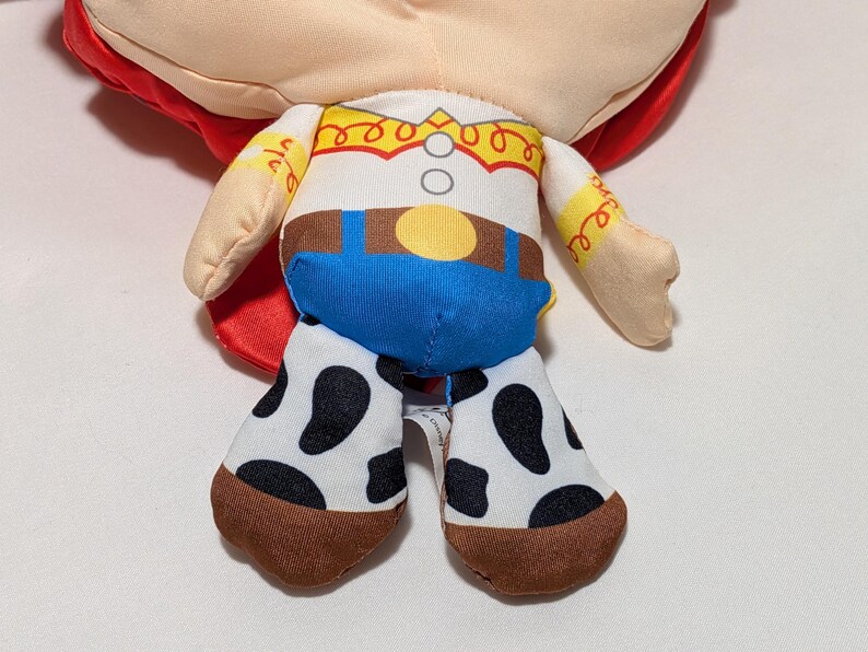 May include: A soft toy doll of a character from the Toy Story franchise. The doll has a white shirt with a yellow yoke, blue trousers, and cow-print chaps. The doll's red hair is visible.