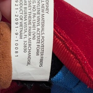 May include: A close-up of a Disney product tag. The tag is white with black text, listing materials, registration information, and the product's origin. The tag is next to a red, blue, and brown plush toy.
