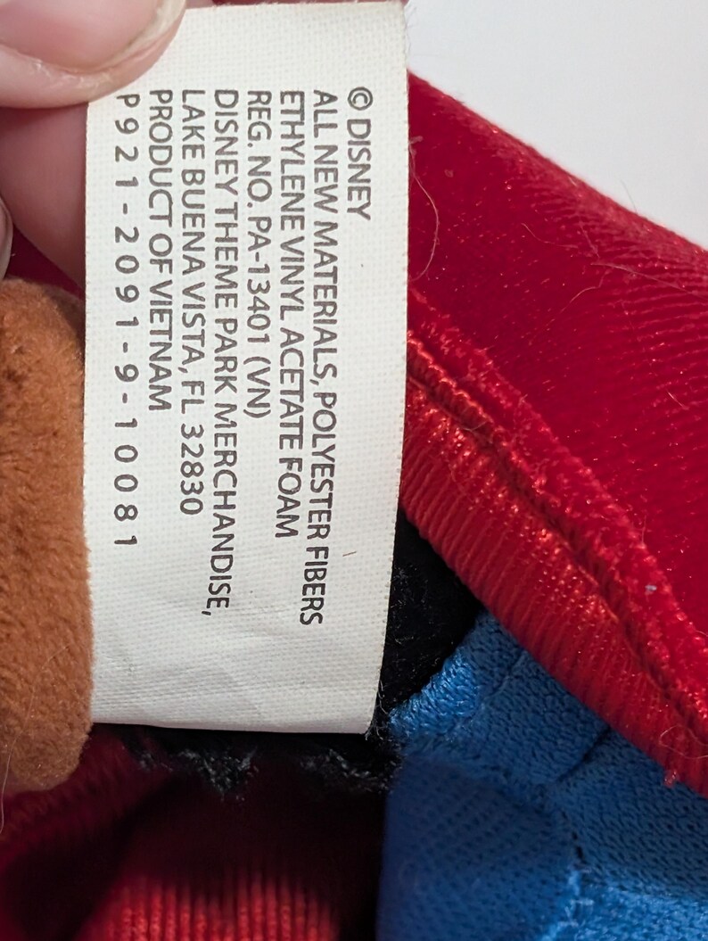 May include: A close-up of a Disney product tag. The tag is white with black text, listing materials, registration information, and the product's origin. The tag is next to a red, blue, and brown plush toy.