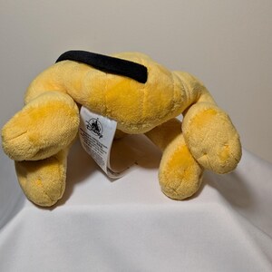 Puede incluir: Un juguete de peluche amarillo con forma de perro y un collar negro. El juguete tiene cuatro patas y una etiqueta con el logotipo de Disney. El juguete est&aacute; sobre una superficie blanca.