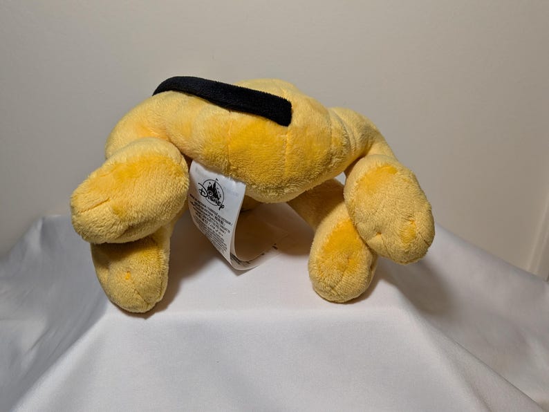 Puede incluir: Un juguete de peluche amarillo con forma de perro y un collar negro. El juguete tiene cuatro patas y una etiqueta con el logotipo de Disney. El juguete est&aacute; sobre una superficie blanca.