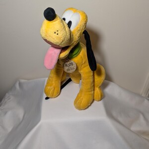 Puede incluir: Un peluche de Pluto, un perro amarillo con orejas negras, una nariz negra y una lengua rosa. Pluto lleva un collar verde y una etiqueta que dice "PLUTO". El juguete est&aacute; sobre una superficie blanca.