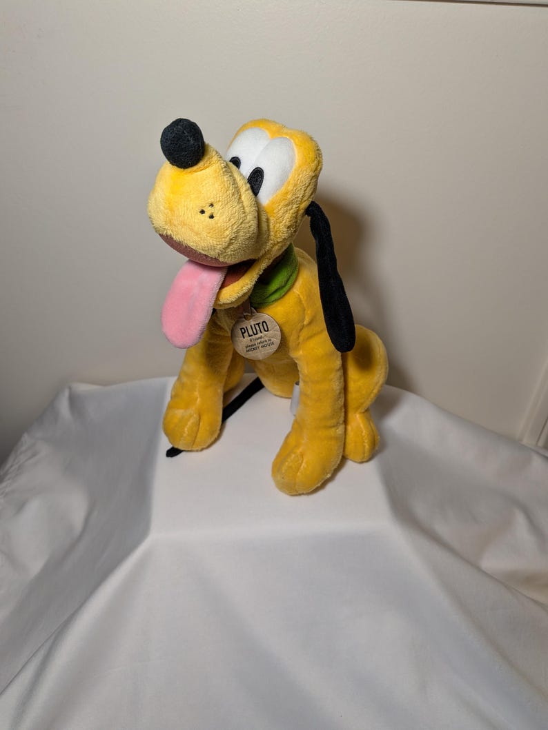 Puede incluir: Un peluche de Pluto, un perro amarillo con orejas negras, una nariz negra y una lengua rosa. Pluto lleva un collar verde y una etiqueta que dice "PLUTO". El juguete est&aacute; sobre una superficie blanca.