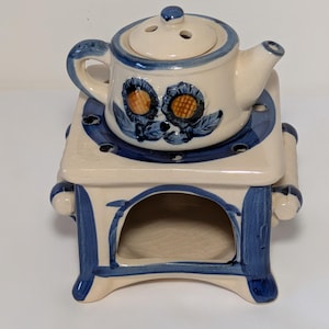 May include: A cream-coloured ceramic teapot warmer set featuring hand-painted blue floral motifs and blue trim. The teapot has a lid with three steam holes, and the square base includes an open arched compartment for a tea light candle.