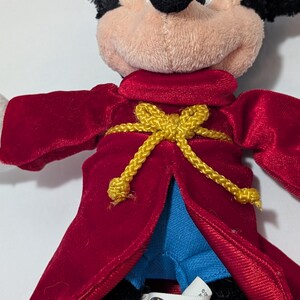 May include: A plush Mickey Mouse doll wearing a red velvet robe with a gold rope belt. The doll has a tan face, black ears, and white gloves. The robe is open, revealing blue fabric underneath.