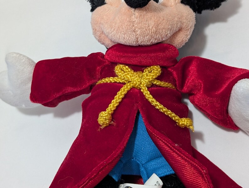 May include: A plush Mickey Mouse doll wearing a red velvet robe with a gold rope belt. The doll has a tan face, black ears, and white gloves. The robe is open, revealing blue fabric underneath.