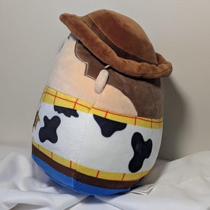 May include: A plush toy shaped like the character Woody from the movie Toy Story. The toy has a brown cowboy hat, a tan face, and a cowhide-patterned vest. The toy is approximately 10 inches tall.