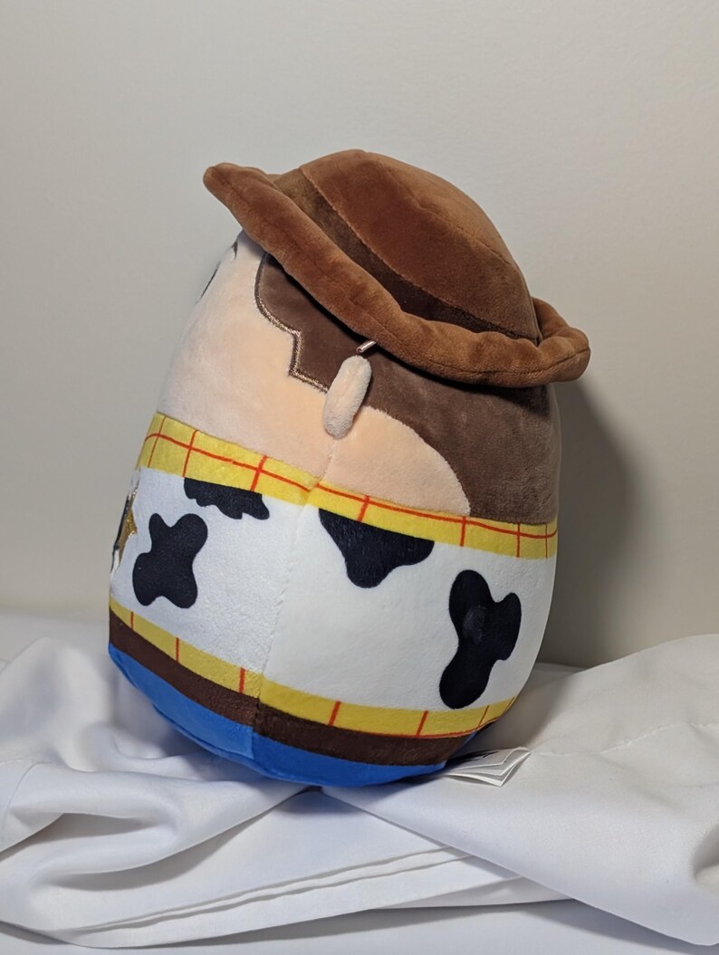 May include: A plush toy shaped like the character Woody from the movie Toy Story. The toy has a brown cowboy hat, a tan face, and a cowhide-patterned vest. The toy is approximately 10 inches tall.