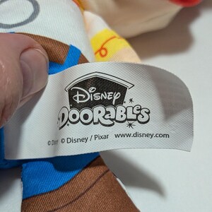 May include: A close-up of a Disney Doorables tag. The tag is white with the Disney Doorables logo and website address. The tag is attached to a plush toy with brown, blue, and white fabric.