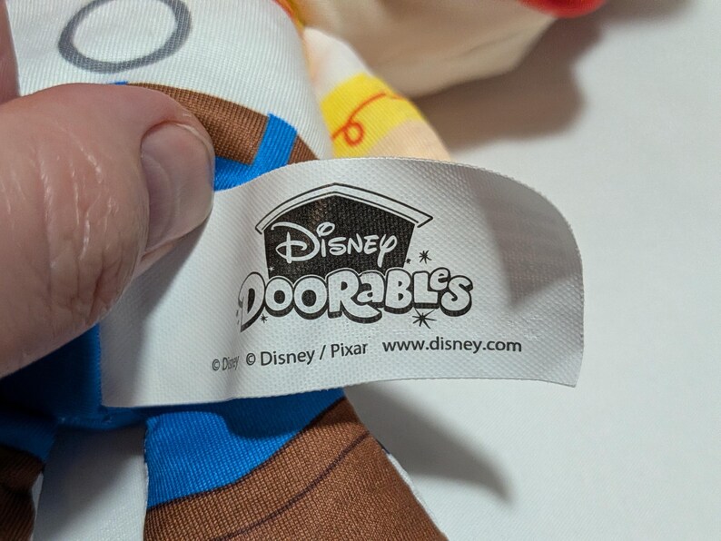 May include: A close-up of a Disney Doorables tag. The tag is white with the Disney Doorables logo and website address. The tag is attached to a plush toy with brown, blue, and white fabric.