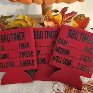 May include: Red BBQ timer can coolers with black text. The text reads "BBQ Timer" with "Rare...1 Beer", "Medium...2 Beers", and "Well Done...3 Beers". The coolers are on a table with fall-themed decorations.
