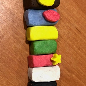May include: A stack of colorful, rectangular clay blocks. The colors include brown with a yellow leaf, blue with a red shape, yellow, green, red with a yellow star, white, and black. The blocks are stacked vertically.