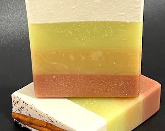 May include: Two bars of handmade soap with a layered design. The top layer is white with brown speckles and a cinnamon stick. Below are layers of yellow, light green, orange, and brown. One bar is on top of the other.