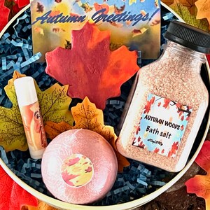 May include: A gift set in a round container with autumn-themed items. Includes a bottle of bath salt, a lip balm, and a bath bomb. The set is decorated with artificial fall leaves and a card that says "Autumn Greetings!"