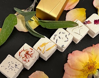 May include: Six small, square bath bombs with various dried flowers and herbs embedded in the surface. The bath bombs are arranged in a row on a black surface.