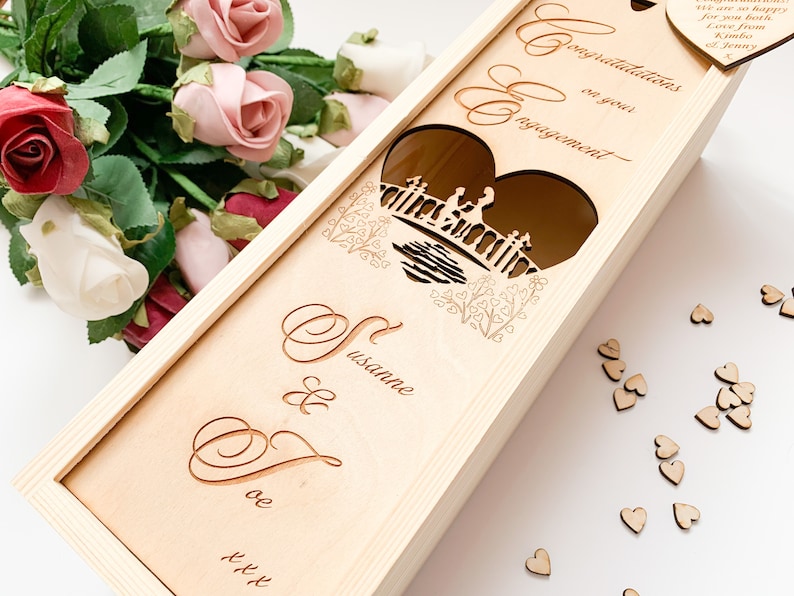 May include: Wooden box with a heart-shaped cutout featuring a couple on a bridge. The box is engraved with the text "Congratulations on your engagement" and the names "Susanne & Dave".