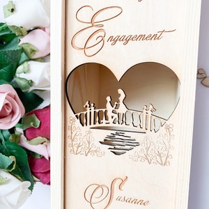 May include: A wooden box with a heart-shaped cutout featuring a couple on a bridge. The box is engraved with the words "Engagement" and "Susanne".
