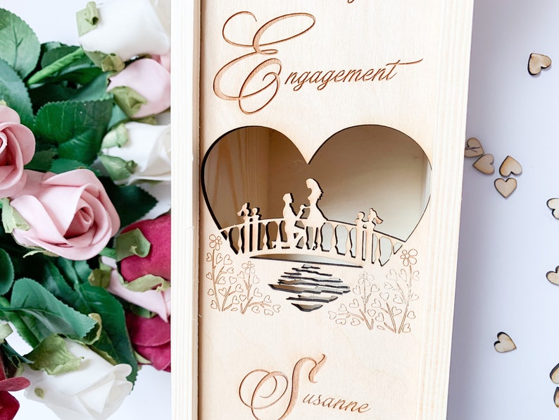 May include: A wooden box with a heart-shaped cutout featuring a couple on a bridge. The box is engraved with the words "Engagement" and "Susanne".