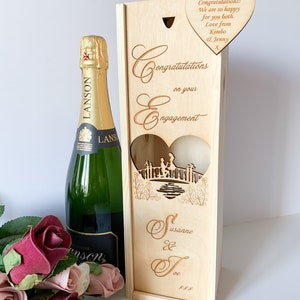 May include: Wooden box with a heart-shaped cutout, designed for a bottle of champagne. The box is engraved with the words "Congratulations on your Engagement" and the names "Susanne & Joe". A separate heart-shaped wooden tag is attached to the box with the message "Congratulations! We are so happy for you both. Love from Kimbo & Jenny X".