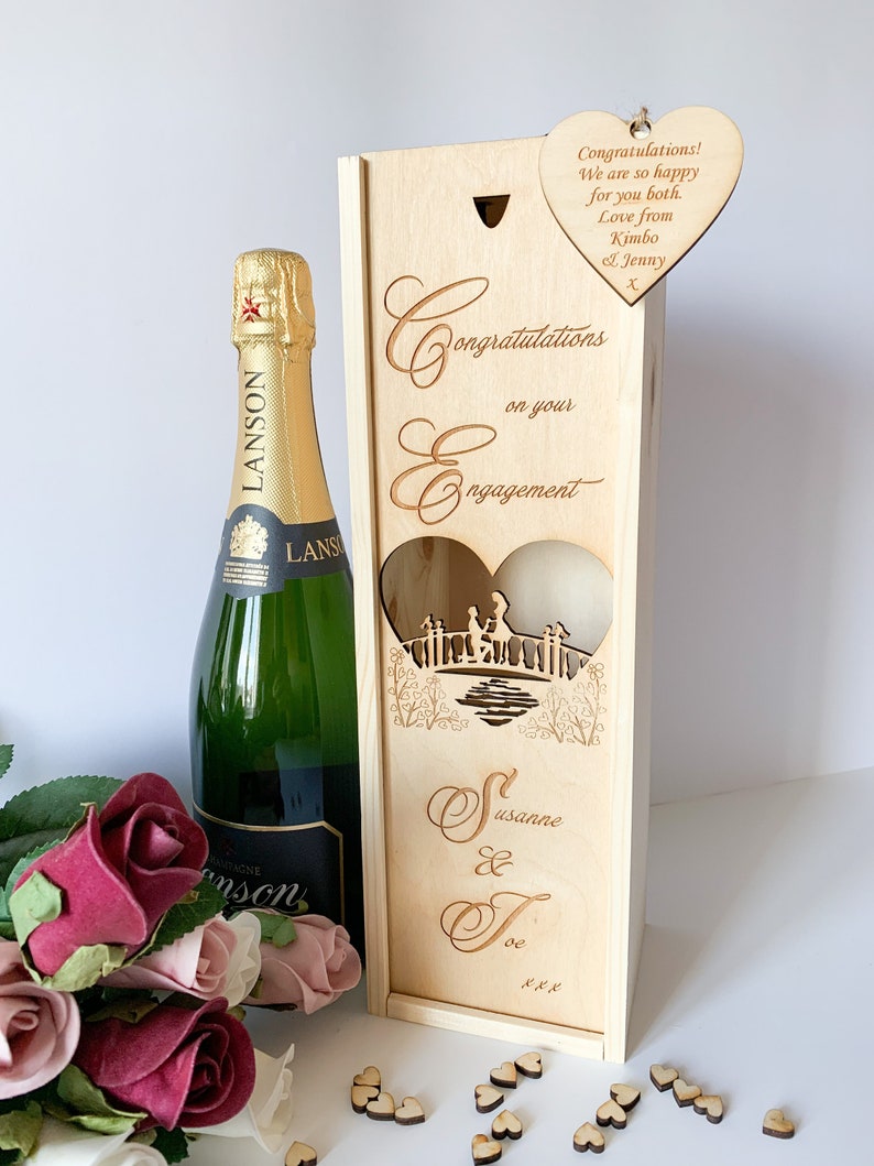May include: Wooden box with a heart-shaped cutout, designed for a bottle of champagne. The box is engraved with the words "Congratulations on your Engagement" and the names "Susanne & Joe". A separate heart-shaped wooden tag is attached to the box with the message "Congratulations! We are so happy for you both. Love from Kimbo & Jenny X".