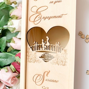 May include: Wooden box with a heart-shaped cutout featuring a couple on a bridge. The box is engraved with the text "Congratulations on your Engagement" and the name "Susanne".
