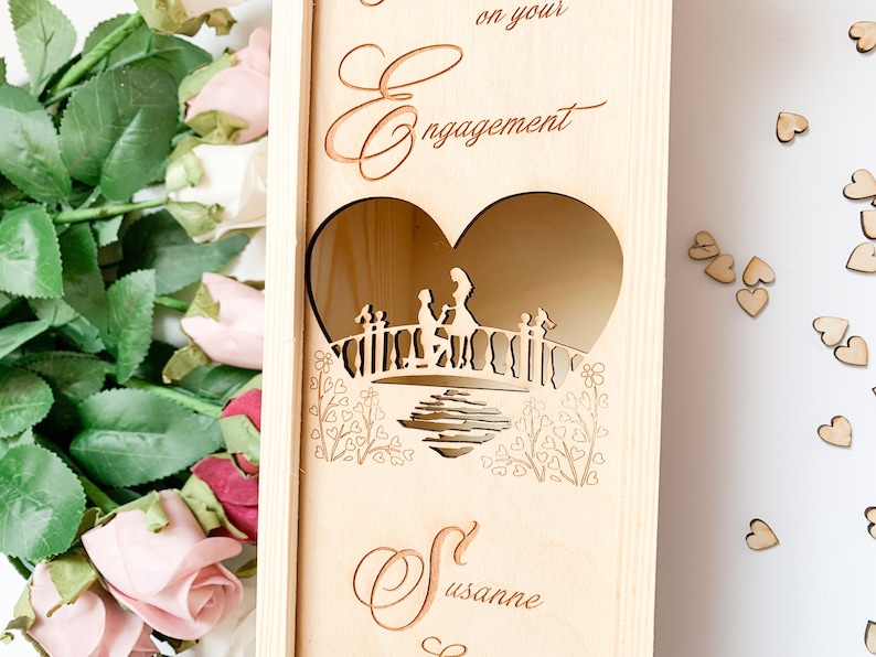 May include: Wooden box with a heart-shaped cutout featuring a couple on a bridge. The box is engraved with the text "Congratulations on your Engagement" and the name "Susanne".