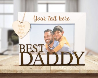 Op de afbeelding: Wit houten fotolijstje met de woorden "Best Daddy" in bruine houten letters. Het lijstje heeft een klein houten hartje dat eraan hangt met de tekst "Lots of love Rohan xx". Het lijstje bevat een foto van een vader en zijn zoon.