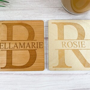 May include: Two wooden coasters with personalized names. The first coaster is brown and features the letter "B" with the name "BELLAMARIE" engraved below. The second coaster is light brown and features the letter "R" with the name "ROSIE" engraved below.