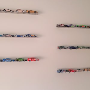 May include: Five rows of toy cars displayed on clear acrylic shelves mounted on a white wall. The cars are a mix of colors, including red, blue, green, yellow, and black.