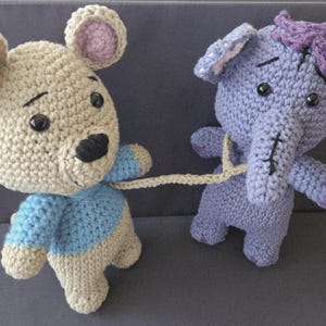 Puede incluir: Dos animales de peluche de ganchillo hechos a mano: un oso beige con una camisa azul y un elefante morado. El oso sostiene una correa atada al elefante. Ambos tienen ojos negros y están hechos de hilo.