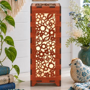Op de afbeelding: Een rechthoekige houten lamp met een bloemenpatroon. De lamp geeft een warme gloed. Het is een decoratief woonaccessoire, geschikt voor een woonkamer of slaapkamer. De lamp is ongeveer 30 cm hoog.