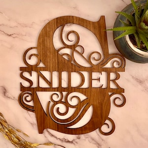 May include: Wooden sign with the surname "SNIDER" cut out in the centre of a large, ornate letter "S". The letter "S" is decorated with intricate scrollwork.