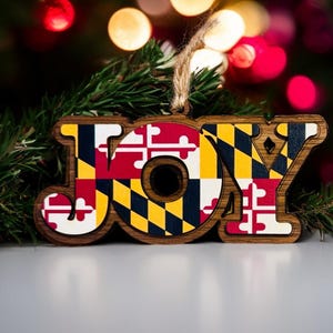 May include: Wooden Christmas ornament spelling "JOY" with the Maryland state flag design. The ornament is brown with the flag's red, white, yellow, and black colors. A natural twine loop is attached for hanging. The background features a Christmas tree and blurred lights.