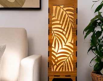 Pode incluir: Uma lâmpada retangular de madeira com um desenho de folhas cortado no painel frontal. A lâmpada é feita de madeira castanha clara e tem uma luz quente e brilhante que emana do interior. A lâmpada é colocada sobre uma mesa castanha escura.