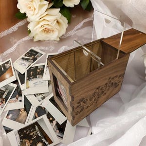 May include: A wooden photo box with a clear acrylic lid and a floral design. The box is open and filled with instant photos.