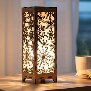 May include: A rectangular wooden lamp with snowflake cutouts. The lamp has a warm, glowing light inside, and is sitting on a wooden surface. The lamp is a decorative item.