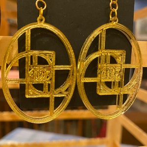 May include: A pair of gold-tone earrings with a geometric design. The earrings are oval-shaped and feature a square pattern with intricate details.