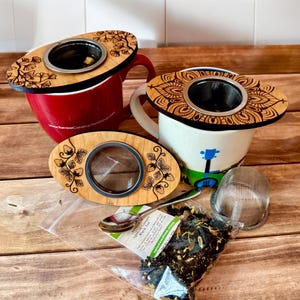 May include: Three wooden tea infusers with different designs. The infusers are oval shaped with a metal mesh strainer in the center. The designs include a floral pattern, a mandala pattern, and a strawberry pattern. The infusers are sitting on a wooden table with a bag of loose leaf tea.