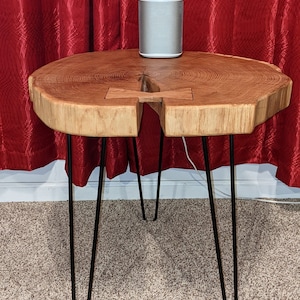 Puede incluir: Una mesa auxiliar de madera de borde vivo con patas de horquilla negras. La mesa tiene un acabado de veta de madera natural y una forma única. Un altavoz plateado y blanco se encuentra encima de la mesa. La mesa mide aproximadamente 76 cm de ancho.