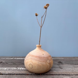 Könnte beinhalten: Eine hellbraune Holzvase mit runder, bauchiger Form. Die Vase ist mit einem einzigen getrockneten Blumenstängel mit drei braunen Blütenköpfen gefüllt.