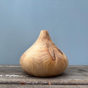 Puede incluir: Un jarrón de madera de color marrón claro con un patrón de veta único y en espiral. El jarrón tiene una forma redondeada y bulbosa y un acabado liso y pulido.