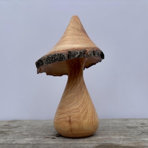 May include: A wooden mushroom sculpture with a light brown cap and stem. The cap has a natural, rough edge. The mushroom is displayed on a wooden surface against a plain background. The sculpture is a decorative item.