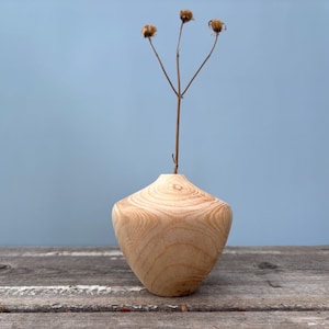 May include: A wooden vase with a natural wood finish and a rounded, bulbous shape. The vase is filled with a single dried flower stem with three brown flower heads.