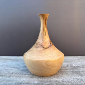 Puede incluir: Un jarrón pequeño de madera de color marrón claro con un patrón de veta de madera natural. El jarrón tiene una base redonda y un cuello estrecho.
