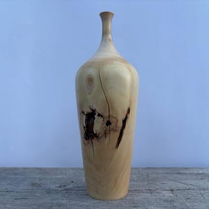 May include: A tall, light-colored wooden vase with a slender neck and a rounded body. The vase features natural wood grain and dark brown knots and markings. The vase stands on a wooden surface against a light blue background.