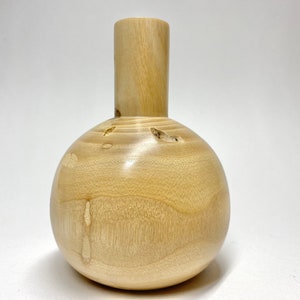 May include: A light wood vase with a round body and a cylindrical neck. The vase is made of natural wood and has a smooth, polished finish.