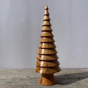 May include: A wooden decorative Christmas tree. The tree is made of light brown wood and has a conical shape with multiple stacked, rounded tiers. The tree sits on a round base and tapers to a point at the top. The wood grain is visible.